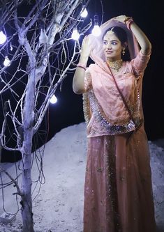 a woman standing in the snow next to a tree with lights on it's branches