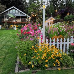 a white picket fence surrounded by flowers and other plants in front of a small house