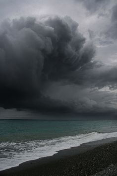 a large storm is coming over the ocean on a cloudy day with black sand and blue water