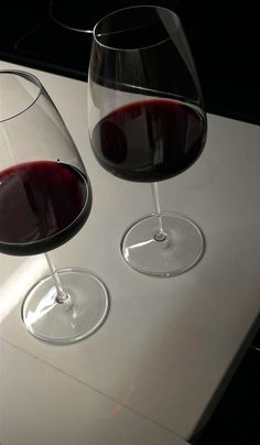 two glasses of red wine are sitting on a white table top, one is half full and the other half empty