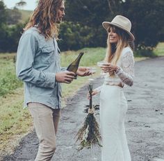 a man and woman standing next to each other on a road holding wine bottles in their hands