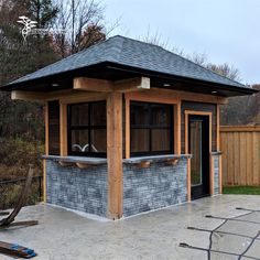 a small stone building with a black roof and wooden posts on the outside, in front of a fenced yard