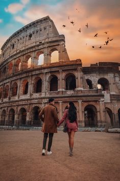 two people standing in front of an old building with birds flying over it at sunset