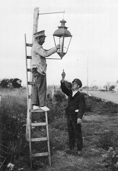 an old black and white photo of two men standing on ladders, one pointing at a lamp