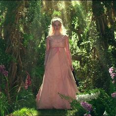 a woman in a pink dress is walking through the woods with flowers and trees around her