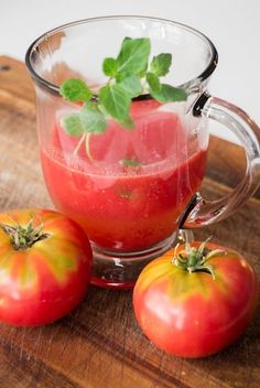 two tomatoes sitting on top of a wooden cutting board next to a glass cup filled with liquid