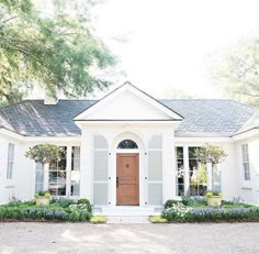 a white house with a brown door and window panes on the front, surrounded by greenery