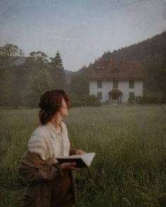 a woman standing in the middle of a field holding a book and looking at a house