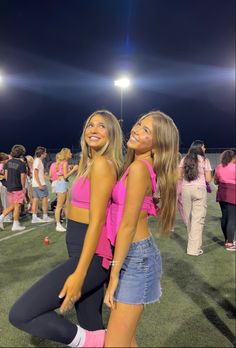 two young women standing on top of a soccer field at night with people in the background