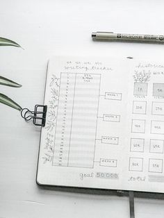 a notebook with a planner, pen and plant next to it on a white surface