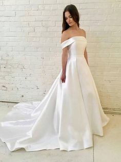 a woman standing in front of a brick wall wearing a white wedding dress with an off the shoulder neckline