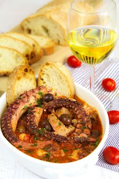 a bowl of soup with bread, tomatoes and olives next to a glass of wine