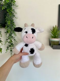 a hand holding a small crocheted cow toy in front of a white wall