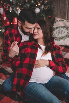 a pregnant couple sitting in front of a christmas tree with their arms around each other