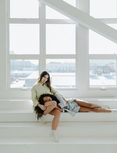 two women sitting on a window sill in front of large windows, one holding the other