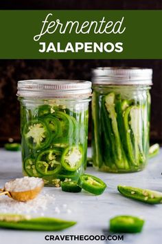 two jars filled with pickles and green peppers next to some salt on the table