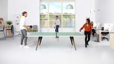 three people are playing ping pong in an office with large windows and desks