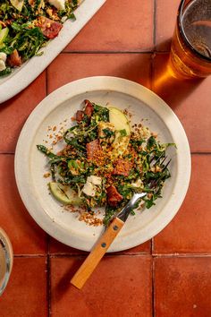 a white plate topped with salad next to a glass of beer on top of a red tile floor