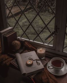 a cup of tea, book and pocket watch sitting on a table next to a window