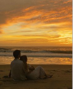 a man and woman sitting on the beach watching the sun go down over the ocean