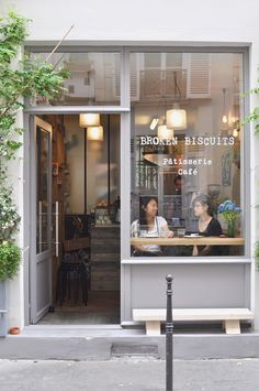 two women sitting at a table in front of a store window with plants growing on the outside