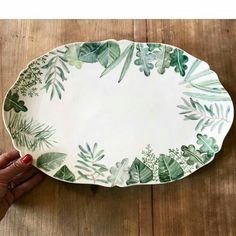 a hand holding a green and white platter with leaves on the rim, against a wooden table