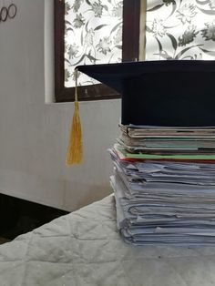 a stack of papers sitting on top of a bed with a graduation cap on top