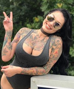 a woman with tattoos on her arms and chest posing for the camera while holding up a peace sign