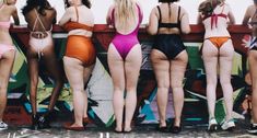 six women in bathing suits are lined up against a graffiti - covered wall with their backs to the camera