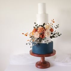 a white and blue cake with flowers on top sitting on a wooden stand in front of a white wall