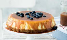 a cheesecake with blueberries and chocolate sauce on a white cake stand next to a book