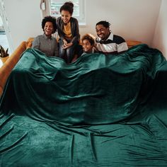 three people are sitting on a bed with a green blanket over them and one person is jumping up into the air