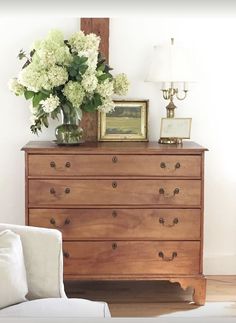 a wooden dresser with flowers on top and a framed picture above it, along with a white chair