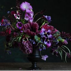 a black vase filled with purple flowers on top of a wooden table in front of a dark background