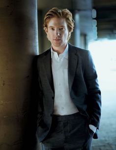 a young man in a suit leaning against a pillar with his hands on his hips