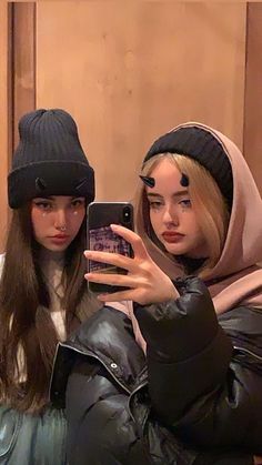 two women wearing beanies taking a selfie in front of a mirror with their cell phone