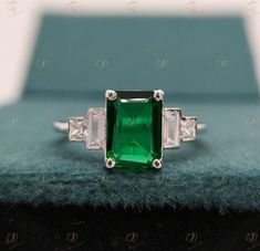 an emerald and diamond three stone ring on top of a blue velvet box with diamonds around it