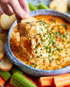 a hand dipping cheese into a bowl of dip surrounded by carrots and celery