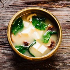 a bowl filled with soup and greens on top of a wooden table next to a spoon