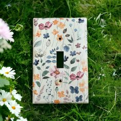 a light switch cover with flowers painted on it in the grass next to daisies