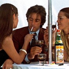 three people sitting at a table drinking wine and talking to each other with an umbrella in the background