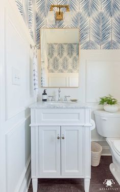 a white bathroom with blue and white wallpaper on the walls, a gold framed mirror above the sink