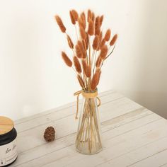 a glass vase filled with dry grass next to a pine cone on a wooden table