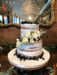 a three tiered cake with white flowers and greenery sits on a table in front of a mirror