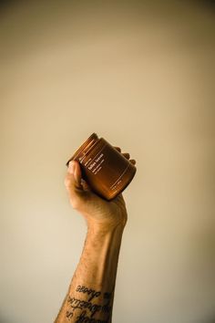 a hand holding up a brown jar with writing on the side and a tattoo on the arm