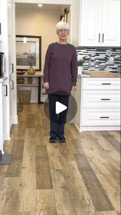 an older woman standing in a kitchen with white cabinets and wood flooring, smiling at the camera