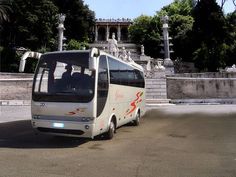 a white bus parked in front of a building with statues on the sides and trees behind it