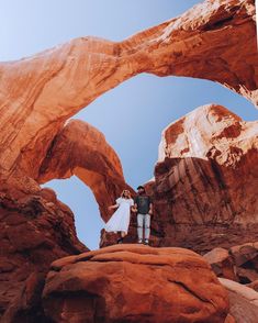 a man and woman standing in front of an arch shaped rock formation with blue sky