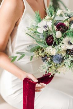 a woman in white dress holding a red ribbon and flower bouquet with greenery on it