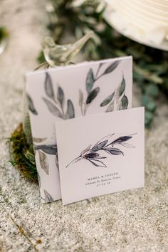 two cards with leaves on them are sitting on the ground next to some flowers and greenery